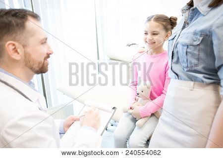 Happy girl with teddybear and her mother talking to doctor while visiting modern clinics