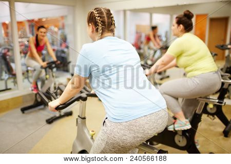 Back view of two young plus-sized women training on bikes in front of mirror at leisure center