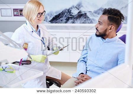 Portrait Of Smiling Female Dentist Talking To Young Man Sitting In Dental Chair During Hygene Consul