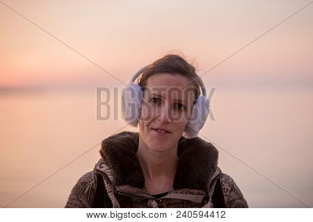 Young Woman Wearing Fluffy Ear Warmers Looking At The Camera Against A Sunset Sky.
