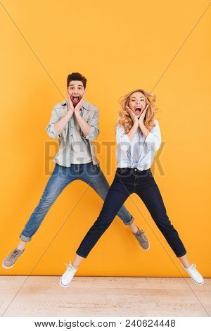 Full length image of happy couple shouting and touching cheeks while jumping with spreading legs isolated over yellow background