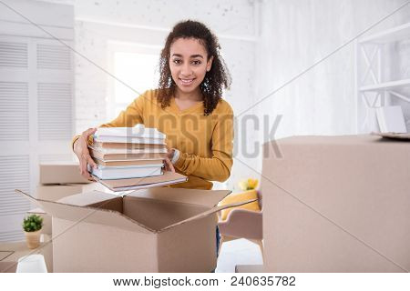 Useful Books. Charming Curly-haired Girl Posing For The Camera And Smiling While Packing A Pile Of B