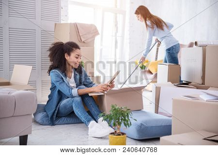 In New Flat. Pleasant Young Girl Cleaning The Floor With A Mop While Her Roommate Sitting On The Flo