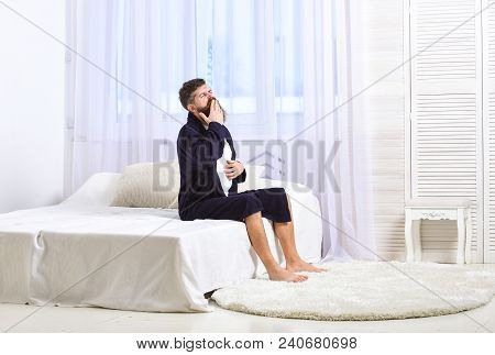 Awakening Concept. Man In Robe Sits On Bed, White Curtains On Background. Macho With Beard And Musta