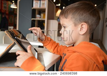 Cute Little Boy Is Sitting In A Cafe At The Table. Plays A Tablet, Watches Cartoons, Reads, Boy Play