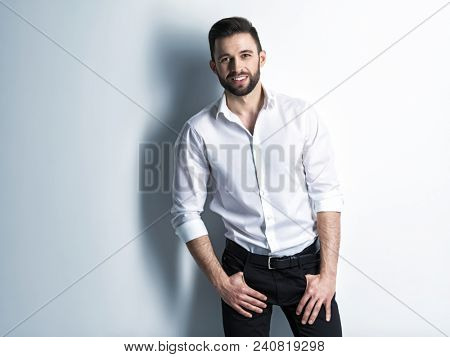 Handsome man with a smile in white shirt and black trousers - posing  at studio. Attractive guy with fashion hairstyle.  Confident man with short beard. Adult stylish boy with brown hair.
