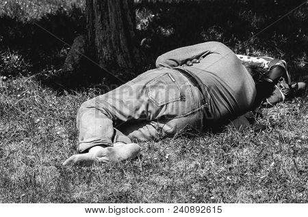 Old Barefoot Homeless Or Refugee Man Sleeping On The Grass In The Park Using His Travel Bag As Pillo