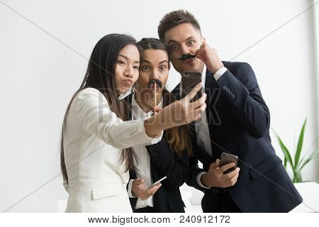 Coworkers Trying Mustache Accessory, Making Silly Faces Posing To Camera While Making Selfie On Smar