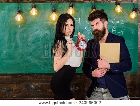 School Rules Concept. Man With Beard Hold Book And Sexy Girl Teacher Holds Alarm Clock, Chalkboard O