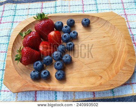Fresh whole, half strawberries and blueberries in wooden bowl on white checkered kitchen towel. Viewed from above.
