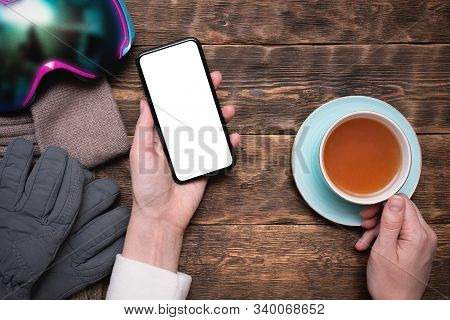 Woman Snowboarder With Blank Screen Mobile Phone Is Drinking A Hot Cup Of Tea On Mountain Resort Caf