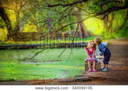 Brother And Sister Walking Near An Artificial Lake Under The Branches Of Trees.