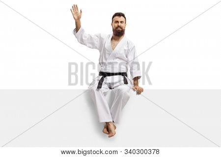 Bearded karate man in kimono with black belt sitting on a panel and waving isolated on white background