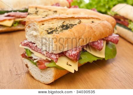 Grupo de sanduíches de baguete longo com alface, legumes, salame, presunto e queijo em uma mesa de madeira