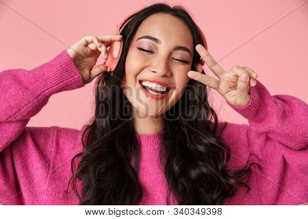 Image of optimistic beautiful asian girl showing peace sign and listening to music with headphones isolated over pink background