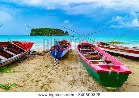 Colorful old wooden fishing boats docked by water on a beautiful beach coast. White sand sea shore landscape on tropical Caribbean island. Holiday weekend/ summer vacation setting at Pagee / Pajee Beach in Port Maria, Jamaica. Cabarita island in the dista