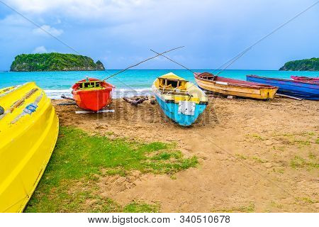 Colorful old wooden fishing boats docked by water on a beautiful beach coast. White sand sea shore landscape on tropical Caribbean island. Holiday weekend/ summer vacation setting at Pagee / Pajee Beach in Port Maria, Jamaica. Cabarita island in the dista