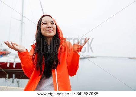 Smiling attractive young asian woman wearing raincoat spending time outdoors walking at the coastland, looking up