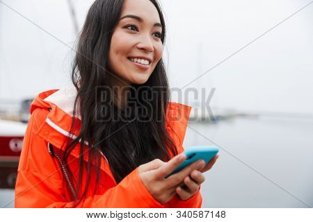 Smiling attractive young asian woman wearing raincoat spending time outdoors walking at the coastland, using mobile phone