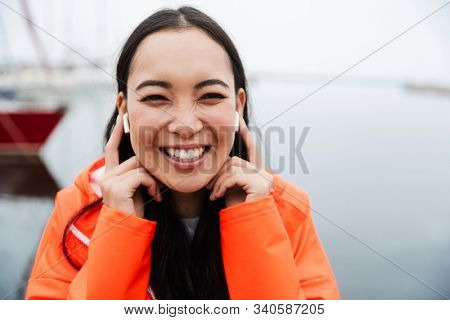 Smiling attractive young asian woman wearing raincoat spending time outdoors walking at the coastland, listening to music with wireless earphones