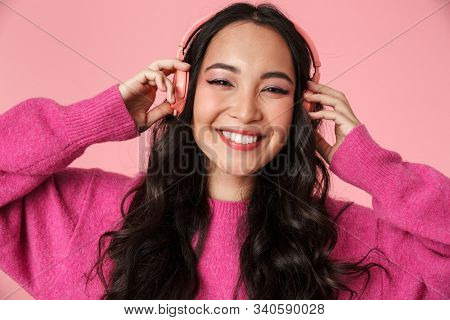 Image of optimistic beautiful asian girl with long dark hair smiling and listening to music with headphones isolated over pink background