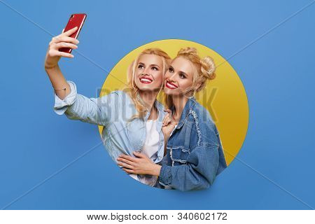 Image Of Two Young Happy Girlfriends Women Standing Over A Blue Background. Twins Peep Out Of A Hole