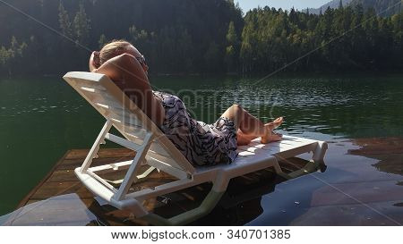 Woman Lie On A Sunbed In Sunglasses And A Boho Silk Shawl. Girl Rest On A Flood Wood Underwater Pier