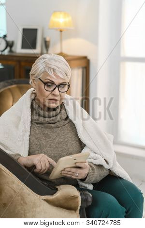 Thougtful Old Lady In Glasses Is Relaxing At Her Comfortable Flat. She Is Sitting On The Leather Arm