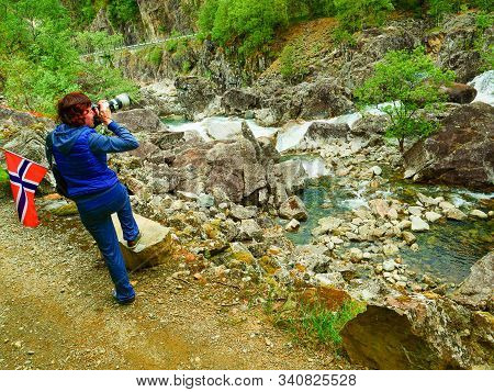 Tourist Woman With Norwegian Flag And Camera Walking Trough Mountains Path In Allmannajuvet Area Sau