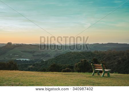 Lone Bench On Green Lawn And Wooded Valley, In A Bucolic Landscape At Sunset Near Bento Goncalves. A