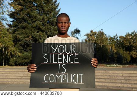 African American Man Holding Sign With Phrase Your Silence Is Complicit Outdoors. Racism Concept