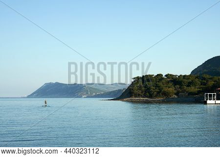 Background Image Of The Sea With A Rocky Winding Shore And A Lone Figure Of A Swimmer On A Board Wit