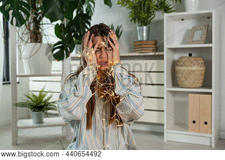 Young Stressed And Frustrated Woman Sitting At Home Having Issue With Tangled Christmas Wire Lights.