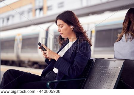 Two Businesswomen Commuting To Work Waiting For Train On Station Platform Looking At Mobile Phones