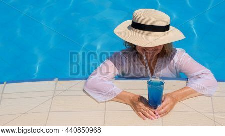 Smiling Senior Woman Relaxing Near The Blue Outdoor Swimming Pool With A Blue Cocktail. People Are E