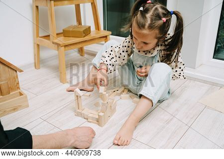 Girl Sits On The Floor In A Room And Plays With Her Dad In A Natral Wood Constructor. Wooden Childre