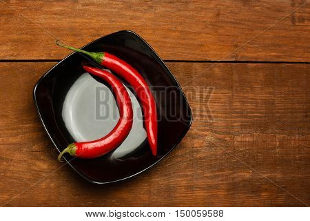 Two red hot cayenne peppers on black ceramic plate standing on brown woden table.