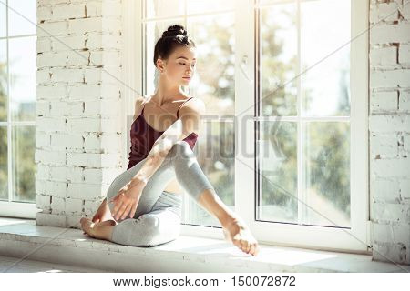 Concentrated on the process. Attractive slim pensive woman sitting near the window and doing physical exercise while having a dance class