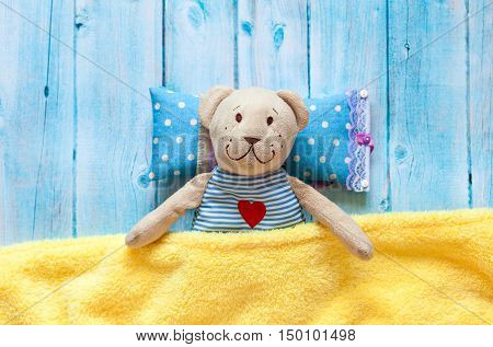 children's soft toy teddy bear in bed with thermometer and pills, take the temperature of a mercury glass thermometer. On a blue wooden background with a yellow blanket. Playing in hospital.