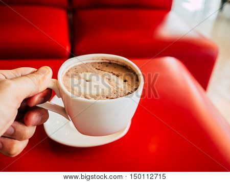 Hand holding white coffee cup on red table.