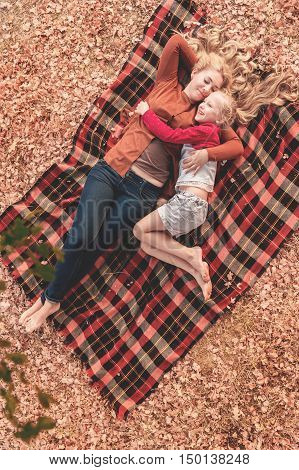 Portrait of two sisters lying under tree on a plaid. Young mom and cute daughter have a good time outsaide. Autumn weather falling leaves.