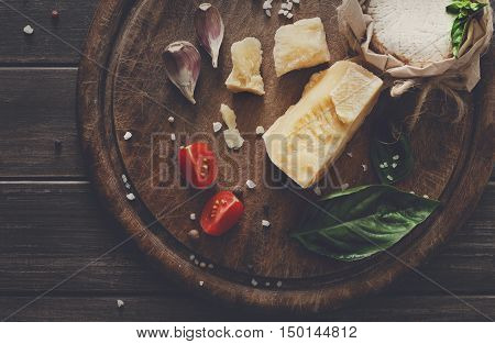 Cheese delikatessen closeup on rustic wood. Wooden desk with parmesan, camembert and brie cuts decorated with garlic, tomato and basil, top view image