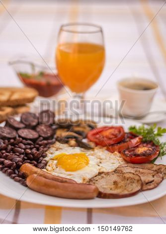Full English fried breakfast with bacon, egg, sausages, black pudding, mushrooms, grilled tomatoes and baked beans. Closeup with toasts, coffee, orange juice and home made  red sauce.