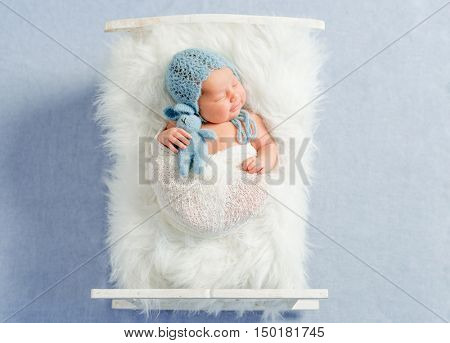dreamy newborn boy holding toy sleeps on little bed covered with white fluffy blanket