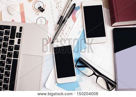Top view of office desktop with two blank smartphones tablet financial report laptop keyboars supplies and other items. Mock up