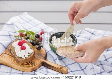 Woman taking organic farming cottage cheese from green bowl. Breakfast: slice of whole wheat bread with Homemade Ricotta cheese served with raspberries and blackberries on wooden board on linen fabric