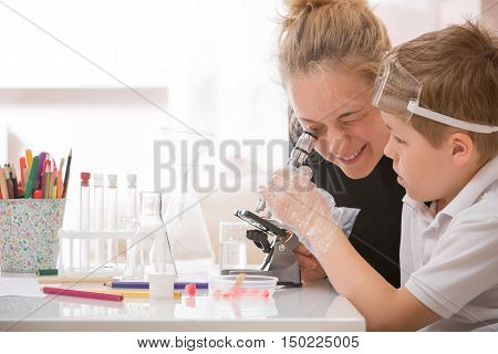 Young mother and elementary school kid boy looking into microscope at home. Family studying samples under the microscope. Science activities with children. Preparing for science lesson.