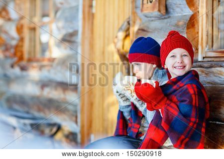 Kids outdoors on beautiful winter day drinking hot chocolate in front of log cabin vacation house