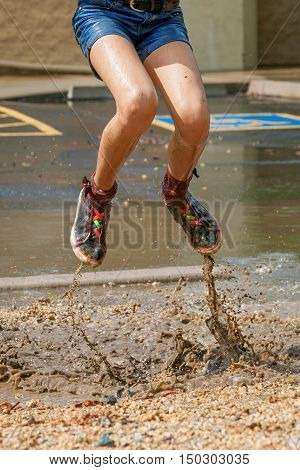 The bottom half of a girl jumping in a muddly puddle in the median of a parking lot. She is wearing jean shorts and colorful shoes and the muddy water is spraying up with her feet.