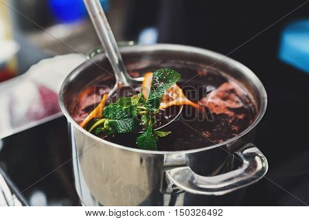 Making mulled wine for sale at country fair. Glintwine with spices, greens, citrus and cloves in big metal stock pot, mixed with soup ladle.